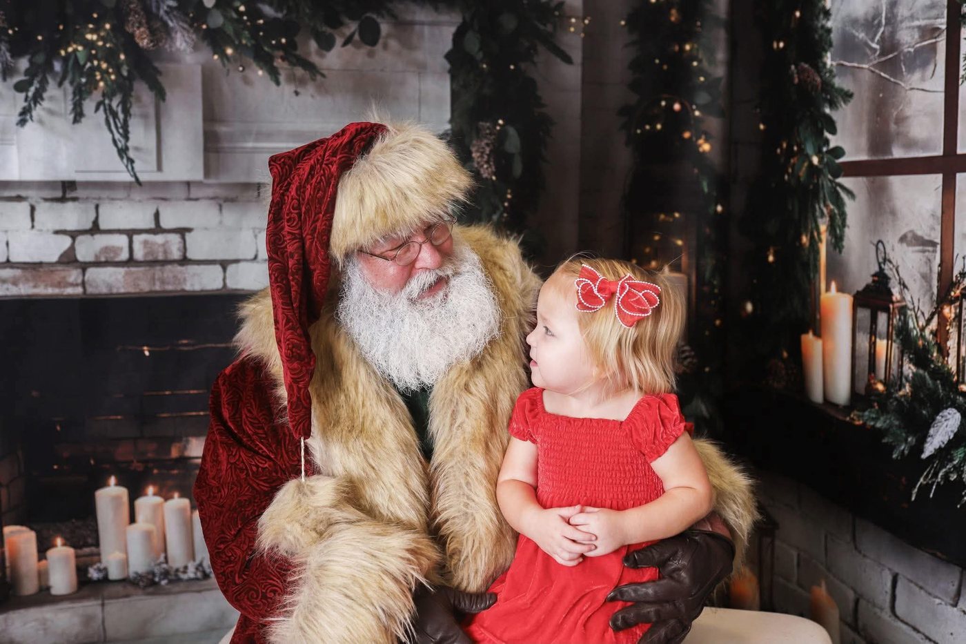 Santa sitting with a small girl in a red dress beside a lit fireplace
